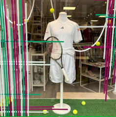 A shop window displaying a mannequin wearing tennis whites and a tennis racquet. Artificial grass is laid on the floor with tennis balls on top.