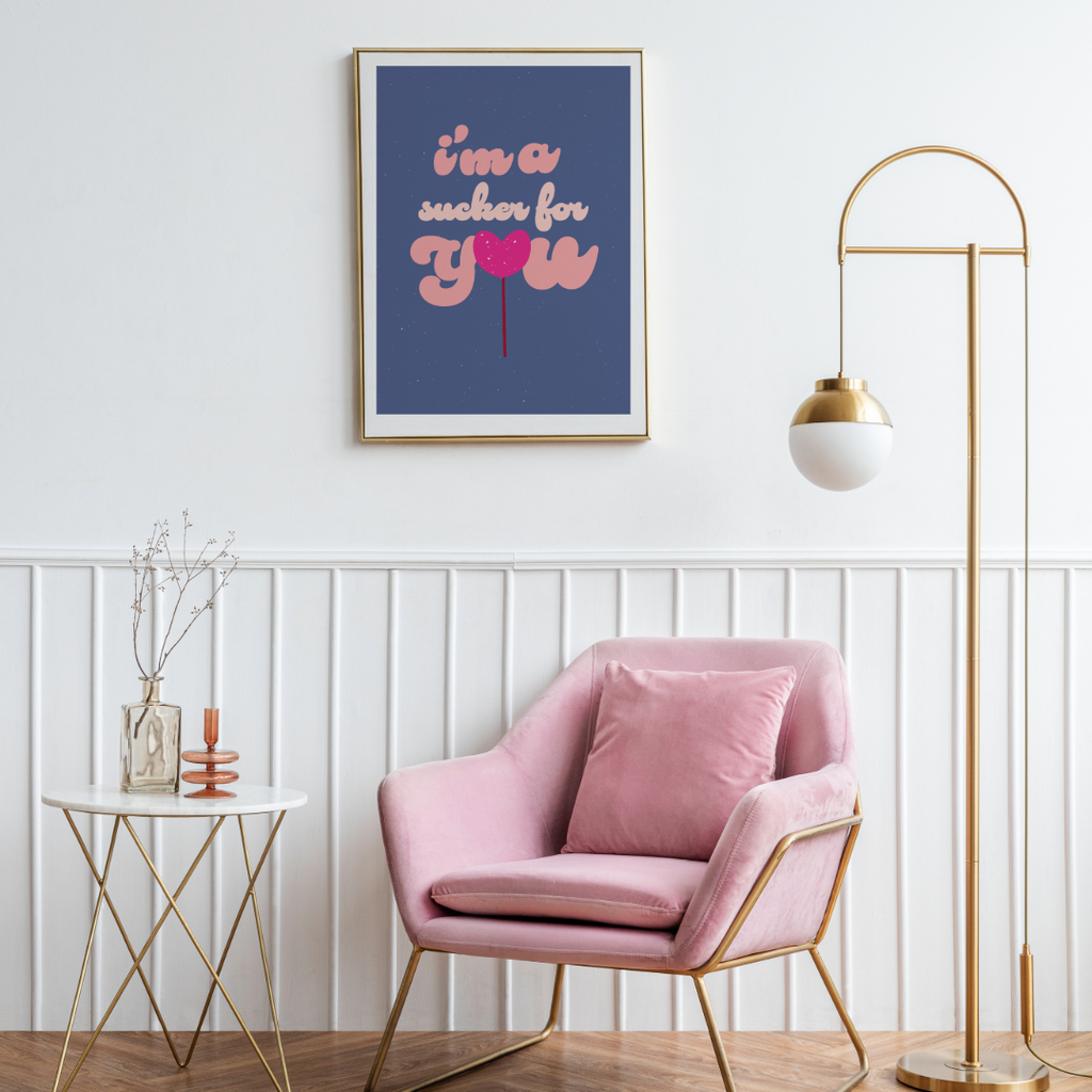 a framed wall print hanging above a pink suede arm chair placed next to a side table with glass décor accessories The text on the wall print says I am a sucker for You