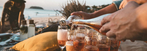 Person pouring champagne into glasses on sandy beach with waves in background