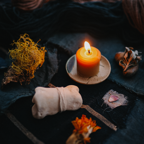 a candle flickers upon an indigo dyed fabric surrounded by lichen, a seashell, a bone wrapped with yarn, and a dried marigold