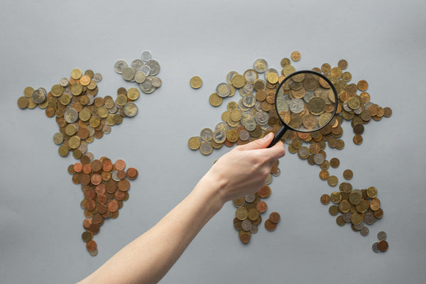 an image of a world map made of coins with a person’s hand holding a magnifying glass over Asia