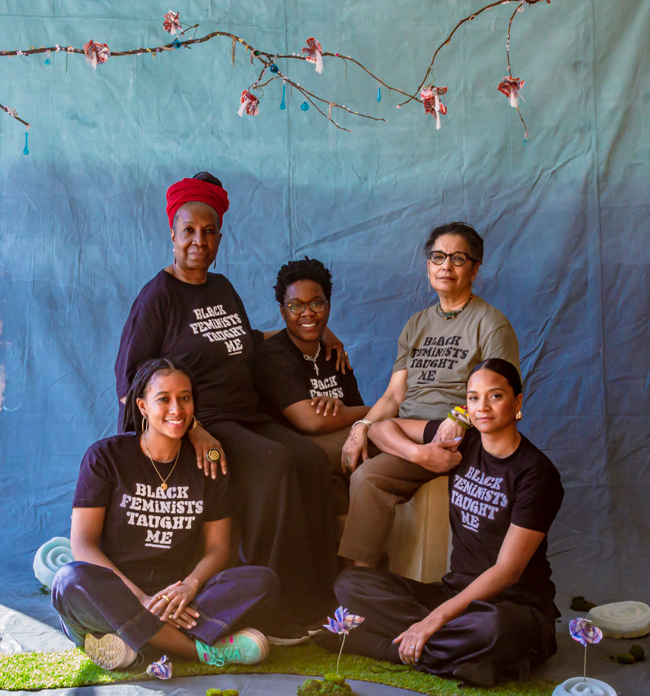 Photo of five people seated in front of a blue backdrop. Each person wears a shirt that says "Black Feminists Taught Me".