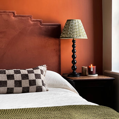 Image of a bed with a brown headboard against an orange wall. There is a lamp with a green shade and and cream bed cover.