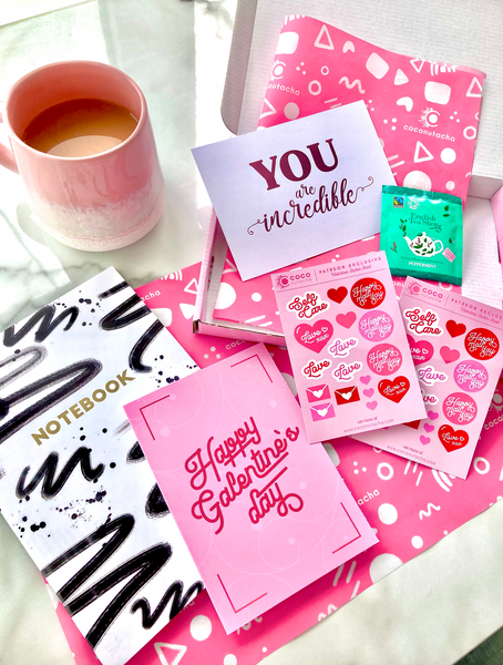 coconutacha patreon goodie box for valentines day featuring a grid, gold foiled notebook, valentines planner stickers, a pink foiled postcard, a galentines day greeting card and a herbal teabag.