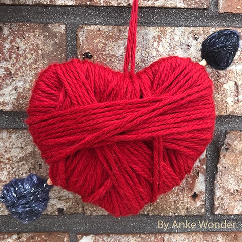 Heart with an arrow out of red and black yarn in front of a brick wall.