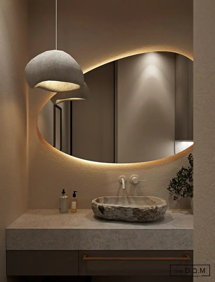 A modern bathroom featuring an artistic oval mirror with light glowing softly around its curved edges, paired with a stone basin, warm pendant lamp, and minimalist countertop accessories