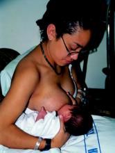 Photo of a woman sitting up, with her baby supported by her right arm, feeding from her right breast.