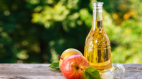 image of apples and apple cider vinegar in a glass bottle on a wooden table with trees in the background