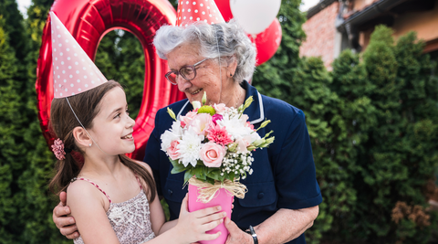 grandma and granddaughter holding a pink and white floral best birthday flowers arrangement and wearing pink party hats