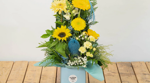 bright yellow floral arrangment of sunflowers, gerberas and others in a blue box on a wooden table