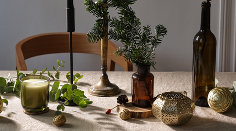 tablescape with gold candles, acorns a brown vase with pine leaves and other greens scattered across a beige tablecloth