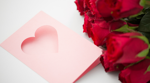 bunch of long stemmed cut roses, red, with a pink card with a heart cut out against a white background
