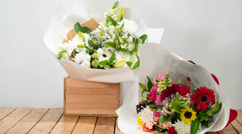 two arrangements, one that's whites and one that is multicoloured including red roses, pink spray roses and yellow sunflowers on a wooden table