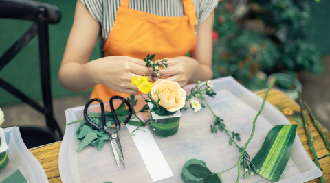 woman in an orange apron sitting at a flower arranging table and arranging table and doing a small arrangement with a rose with scissors sitting next to her