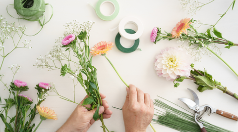 white table with two hands holding gerberas and baby's breath with other blooms and flower arranging tools around