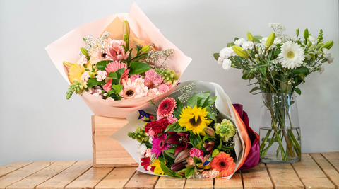image of three flower farm bouquets, one in a pink box, one wrapped and lying on its side and one all white arrangement in a glass vase all on a wooden table