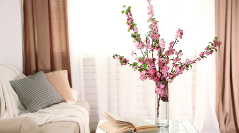 pink climbing blooms, naturally arranged in a clear vase in front of a curtained window next to a couch with cushions and a blanket and with an open book next to it demonstrating flower trends like a floral centred home
