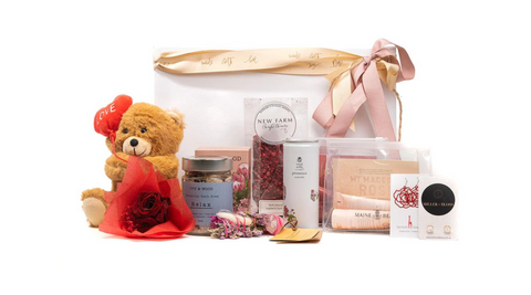 stuffed bear with love heart balloon, some sweets, bath salts, a small bunch of flowers brisbane and some earrings with a gold and pink ribbon behind on a white background