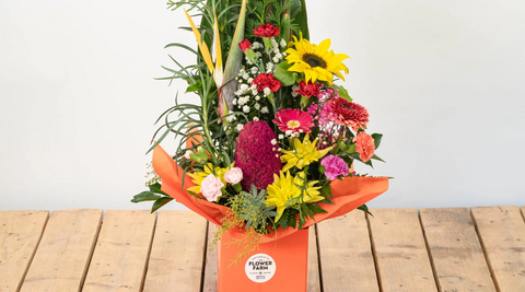bright arrangement of yellow, pink flowers for baby boy with greenery in orange box on a wood table