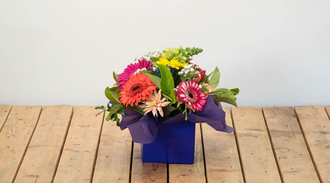 image of a bunch of multi-coloured gerberas, pink, orange, yellow and more, in a blue square container sitting on a wooden slatted table