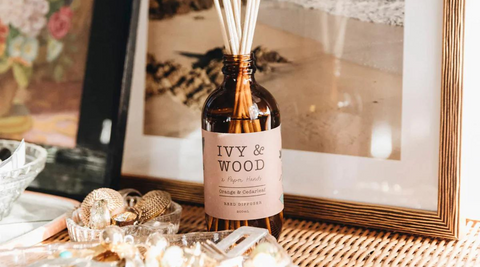 brown bottle of ivy & wood reed diffuser against a neutral framed photo surrounded by glass boxes with jewellery
