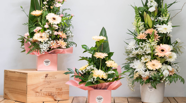 three local flowers arrangements in pale pastel colours one sitting on top of a box and all three arranged across a wooden table