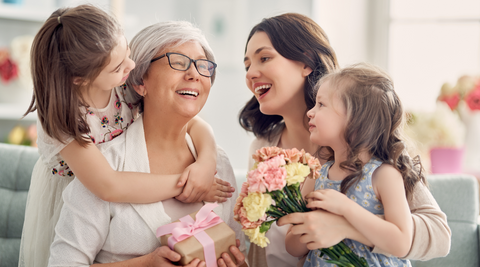 image of a grandmother, mother and two little girls smiling and laughing together, with the grandmother holding a wrapped present and the mom holding a bouquet of chrysanthemums for mothers day card ideas