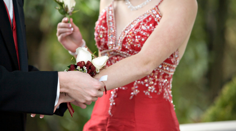girl in red formal dress having a red flower corsage placed on her wrist by her date in a black and red tux while she holds a boutonnière