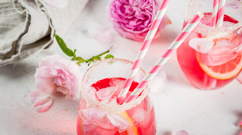 two glasses with pink strawberry rose flower cocktails, with pink roses on a white table cloth and pink and white straws