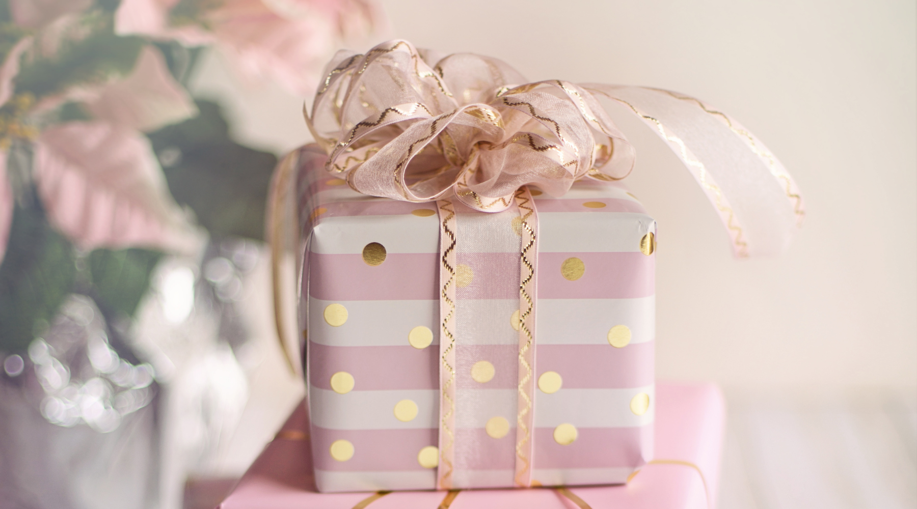 Image of gift wrapped in pink and white paper with a gold bow in front of a pink flower arrangement