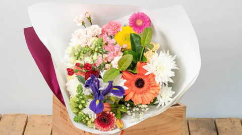 image of mixed bouquet of multi coloured flowers featuring gerberas and mums wrapped in white craft paper on a wooden table for unique mothers day gifts and flowers