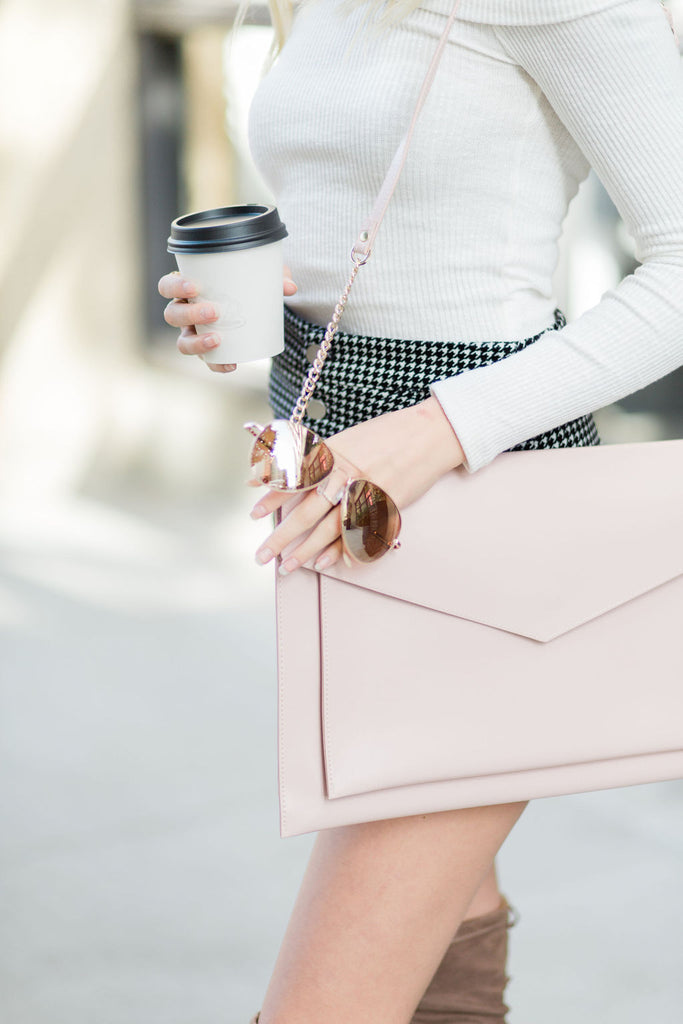 A woman is seen from the shoulders down. She is wearing a long sleeve white shirt and a black and white skirt. She has a coffee and sunglasses in her right hand and over her left shoulder she is wearing a blush pink Hepburn. The thin bag has a thin strap with gold chain accents.