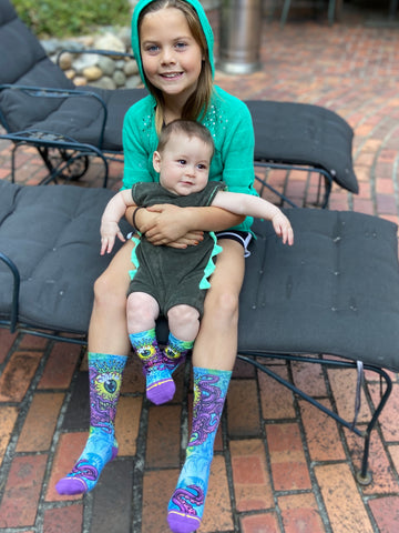 Girl holding baby while wearing a green hoodie outside and Jimbo Phillip's graphic art crew socks for kids and babies.