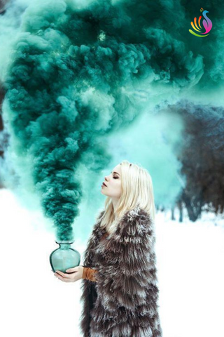 Young blonde woman standing in the snow holding a glass ball with teal green smoke bomb lit inside