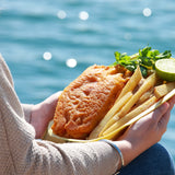 Crispy golden fish paired with a side of fries, garnished with a sprig of cilantro and a slice of lime, being held by someone in a cozy sweater against the shimmering backdrop of sunlit waters.