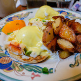 Delectable seafood Eggs Benedict topped with creamy hollandaise sauce, accompanied by crispy roasted potatoes, all served on a vibrant, intricately designed plate, with a slice of fresh orange adding a splash of color.