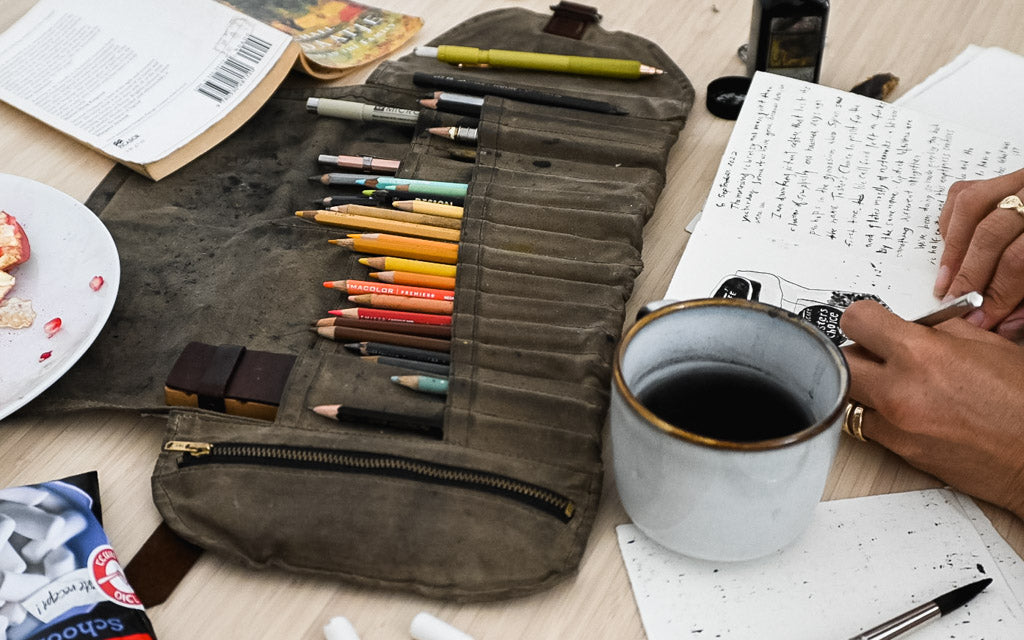 Drawing in a journal with supplies in a brown Sendak Artist Roll filled with colored pencils, fountain pens, and paint brushes