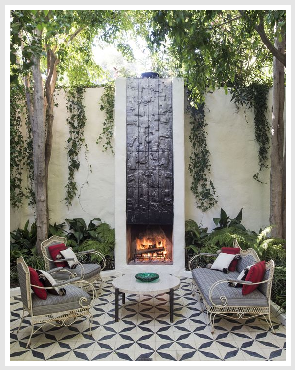 Outdoor fireplace with a metal chimney, 2 elegant chairs, a sofa, and a coffee table, surrounded by a stone patio.