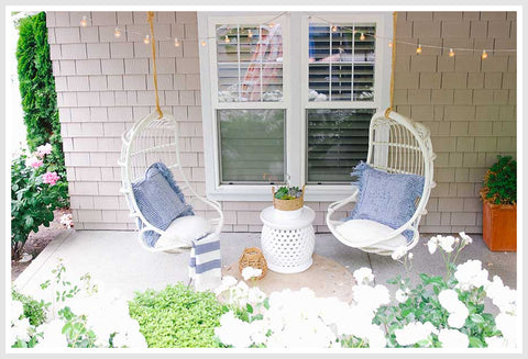 2 white rattan hanging chairs hanged on a patio. White accent table between them.
