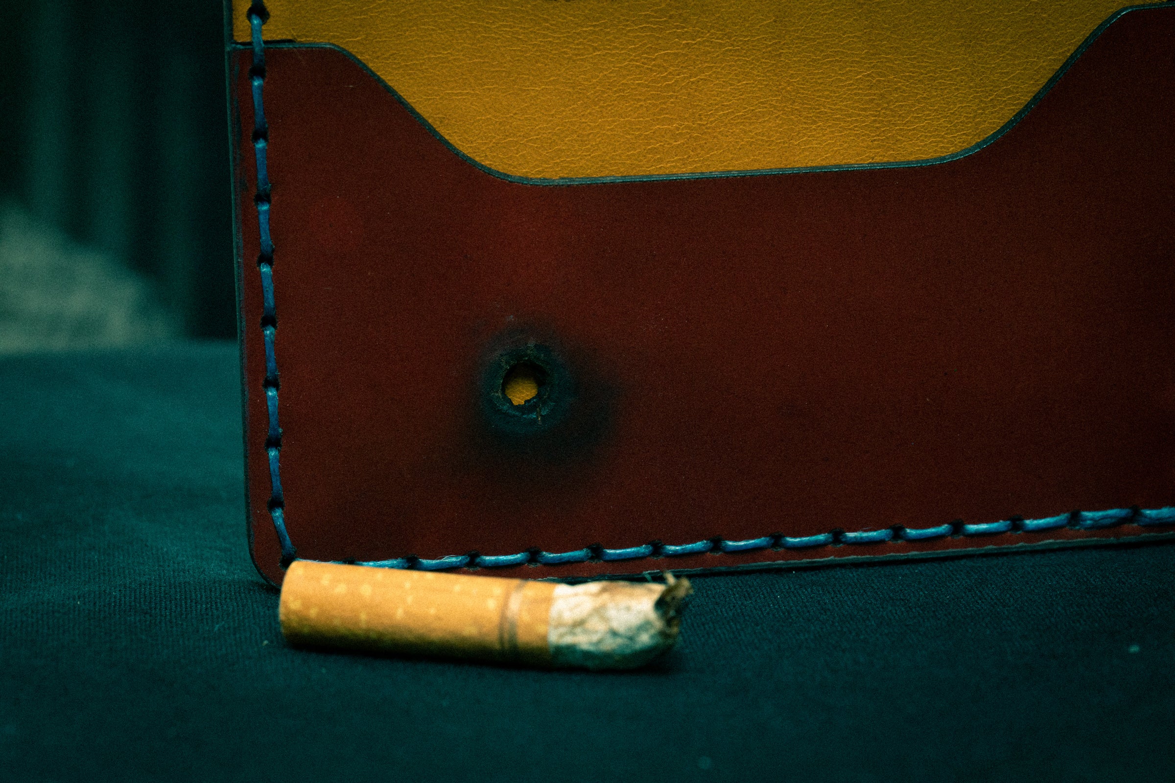 A close up of a maroon pocket of a wallet displays a circular burn hole mark from a cigarette that sits in front
