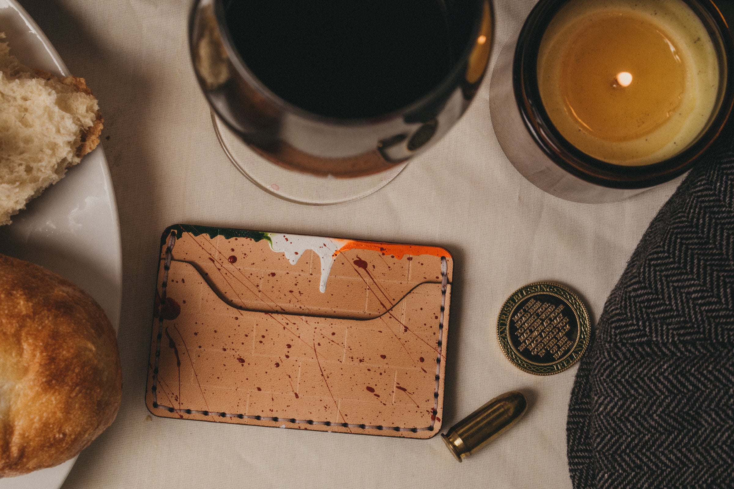 A light tan colored slim wallet sits on top of a table with splatter of red with dripping Irish flag paint across top. In the fore ground a bullet and glass of wine, bread, an unknown coin ,and a candle can be seen.