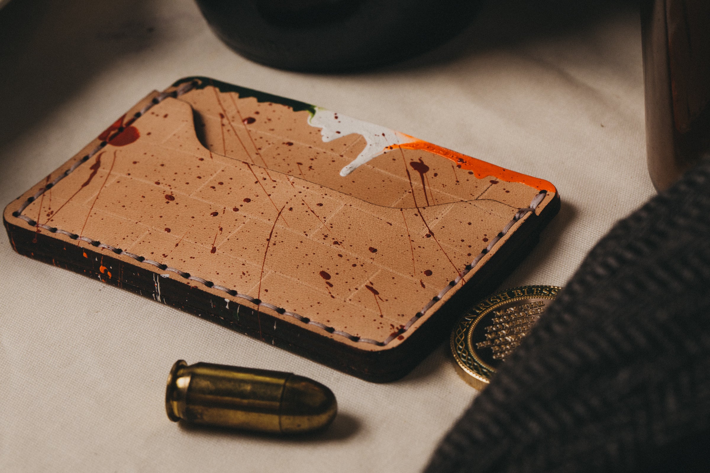 A light tan colored slim wallet sits on top of a table with splatter of red with dripping Irish flag paint across top. In the fore ground a bullet, an unknown coin, and the edge of an older hat can be seen.