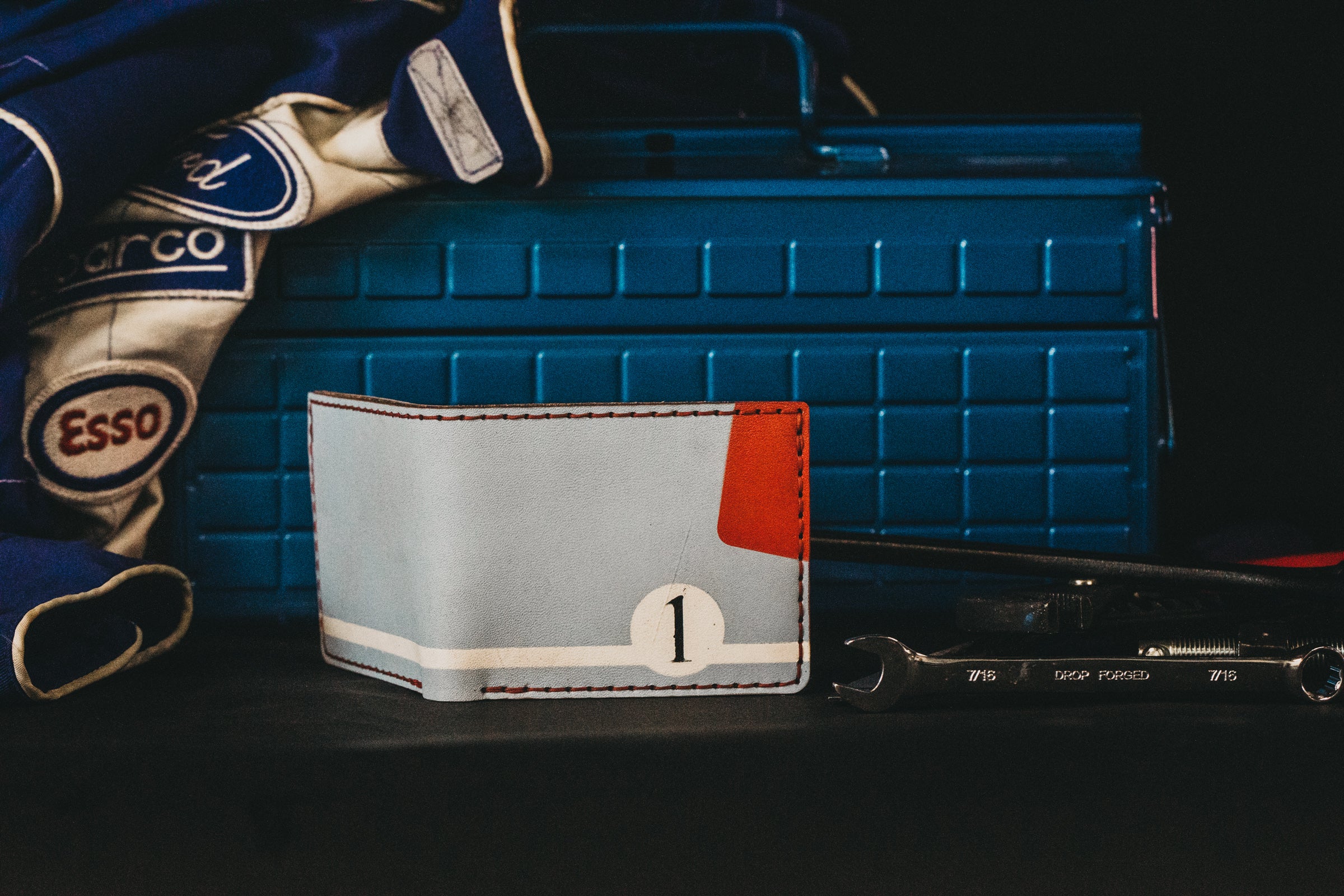 A light blue mechanic theme with red accents wallet sits in front of a darker blue metal tool box with a jacket draped on top of it. 