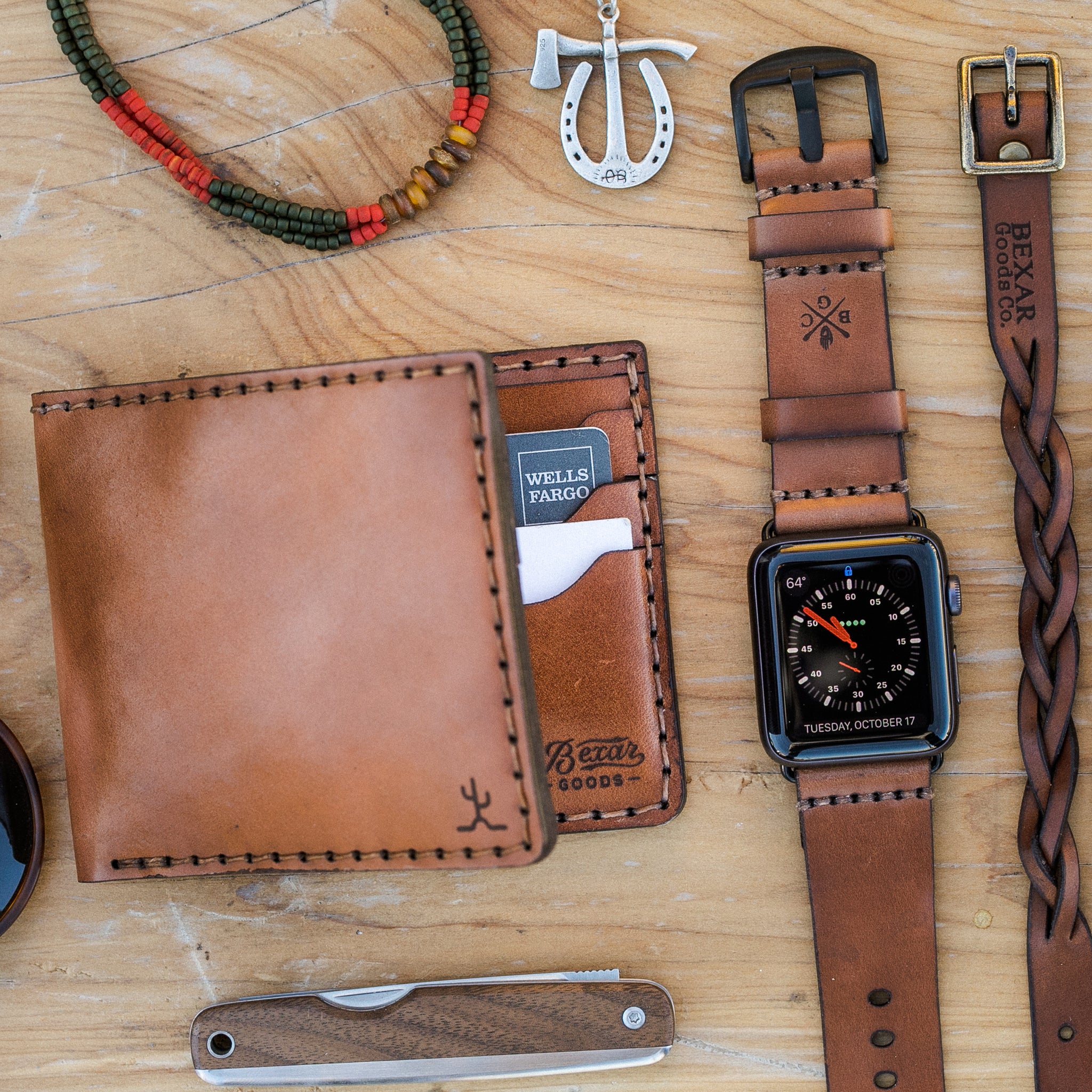 Leather accessories sit ontop of a wood table top. Leather Bracelet, Apple Watch Strap and bifold wallet. As well as a metal and wood pocket knife. As well as a beaded bracelet and horseshoe and hatchet silver necklace on a leather cord.