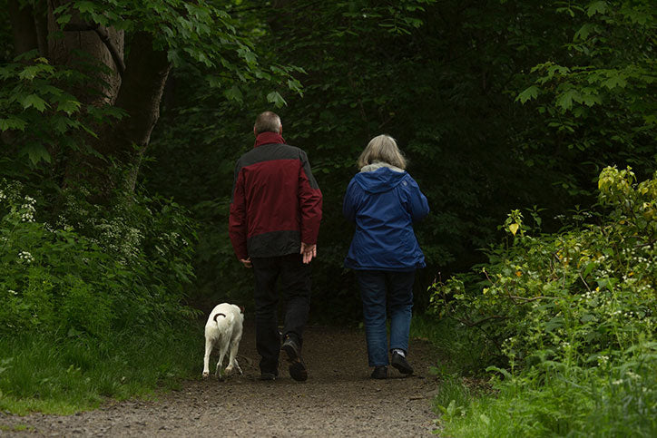 A man in a red jacket and woman in a blue jacket walk along a trail in a forest alongside a white dog