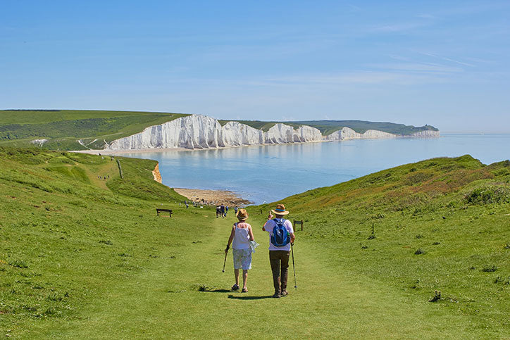 An older man and woman carrying canes walk along a green grassy shore toward a beach and blue body of water with white cliffs in the background
