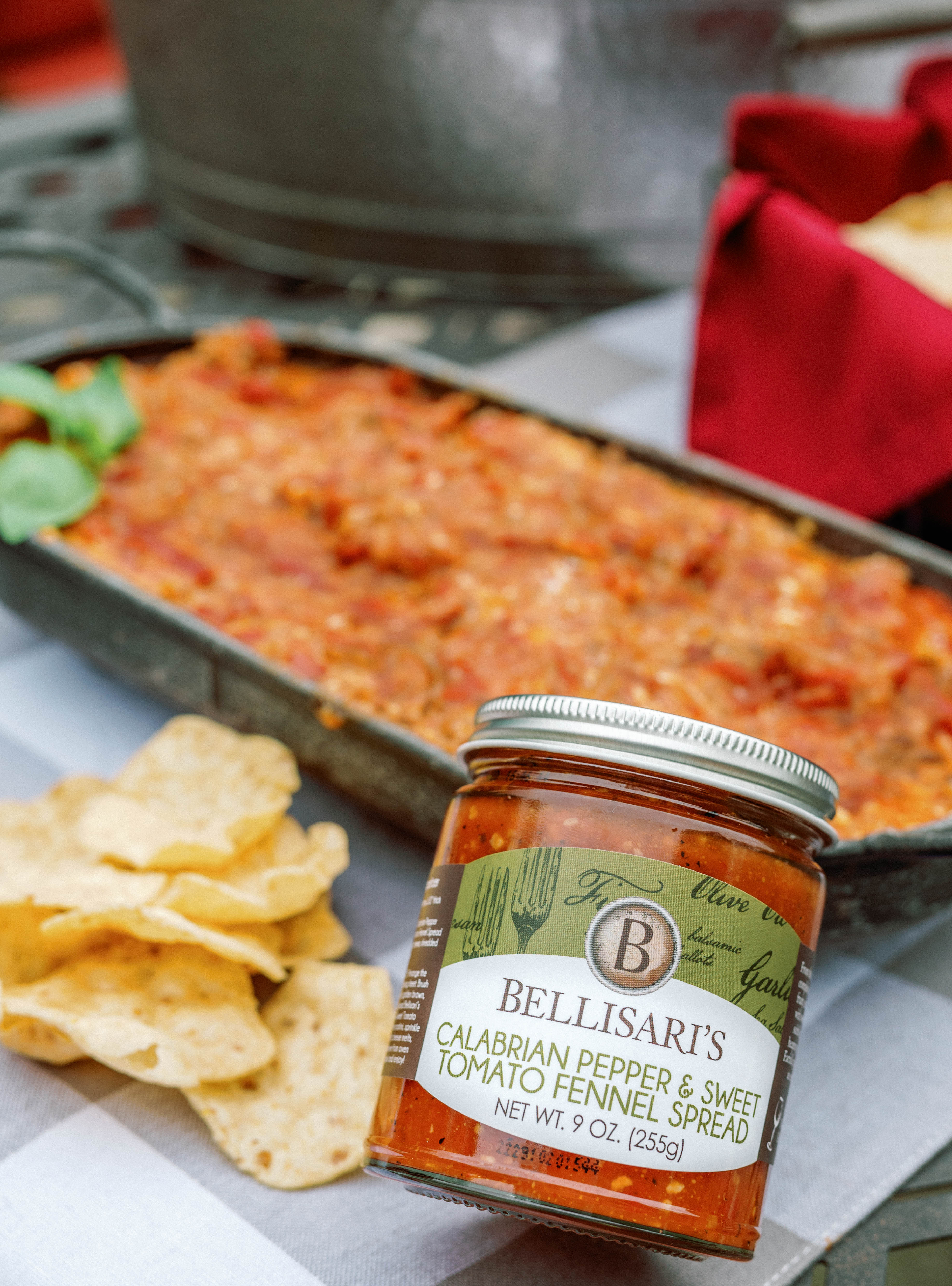 Bellisari's Calabrian Pepper & Sweet Tomato Fennel Spread next to lasagna dip and chips on a picnic table. 