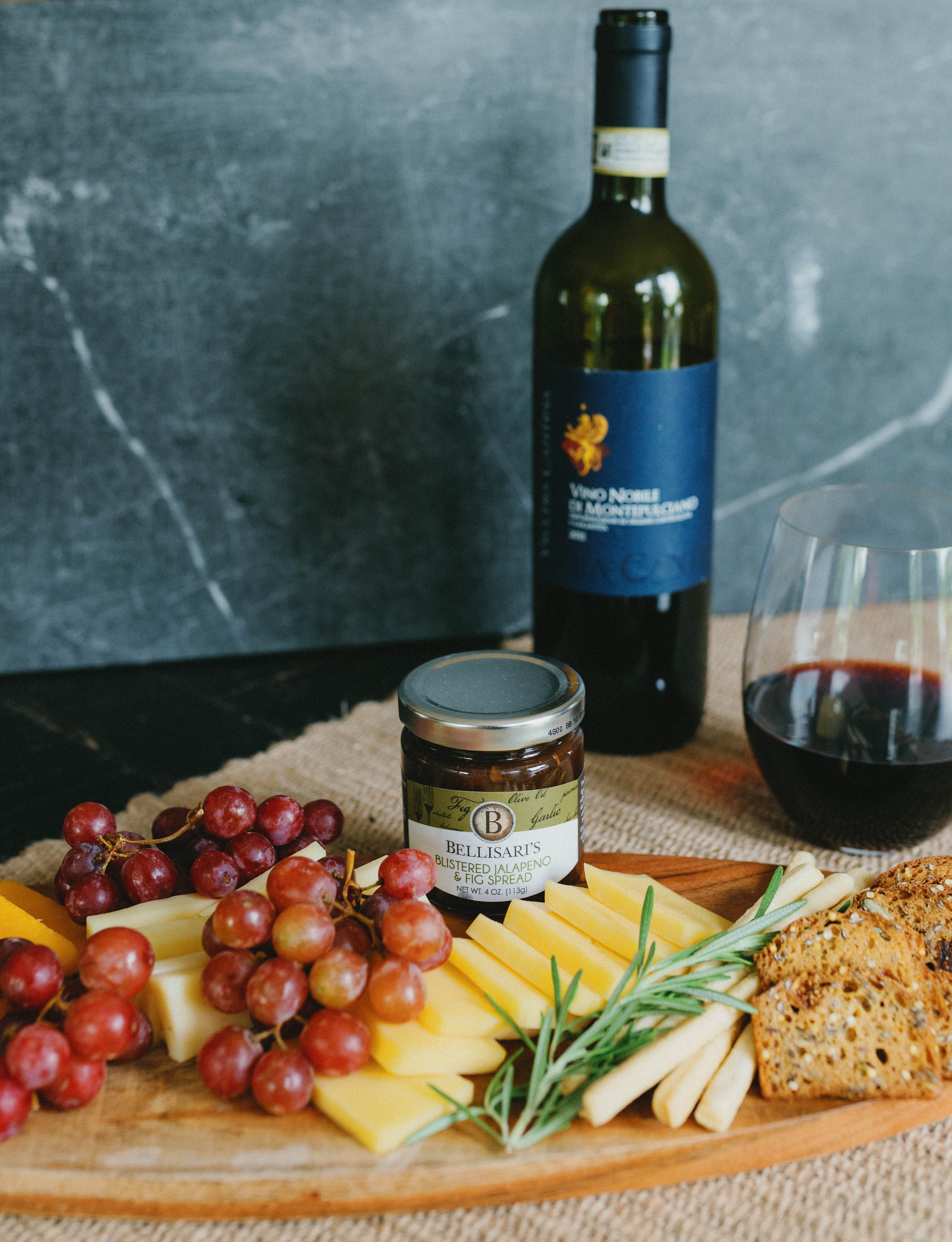 A bottle of wine with a Bellisari's jar and a charcuterie board with cheese and grapes. 