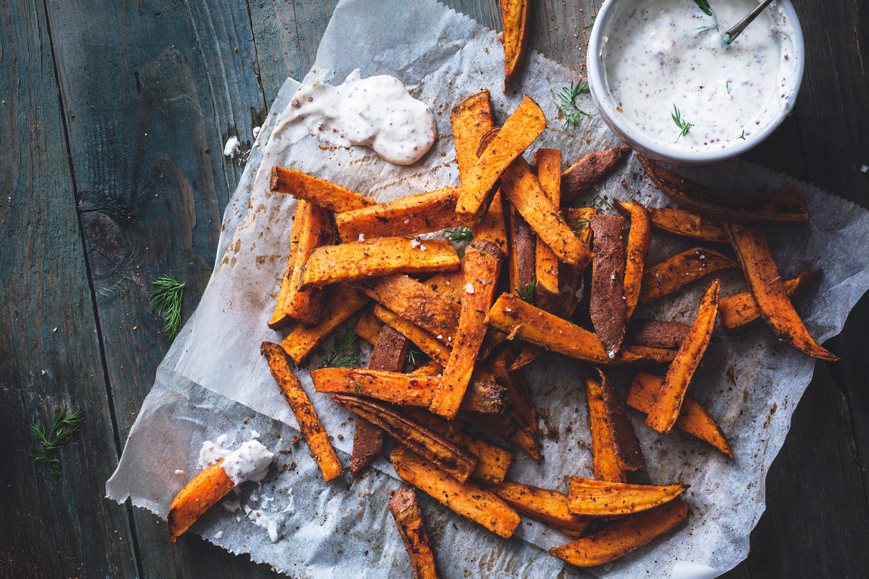 Sweet potato fries on parchment paper with a dill dipping sauce next to them. A dark wood is the background and creates a moody feel to the image.