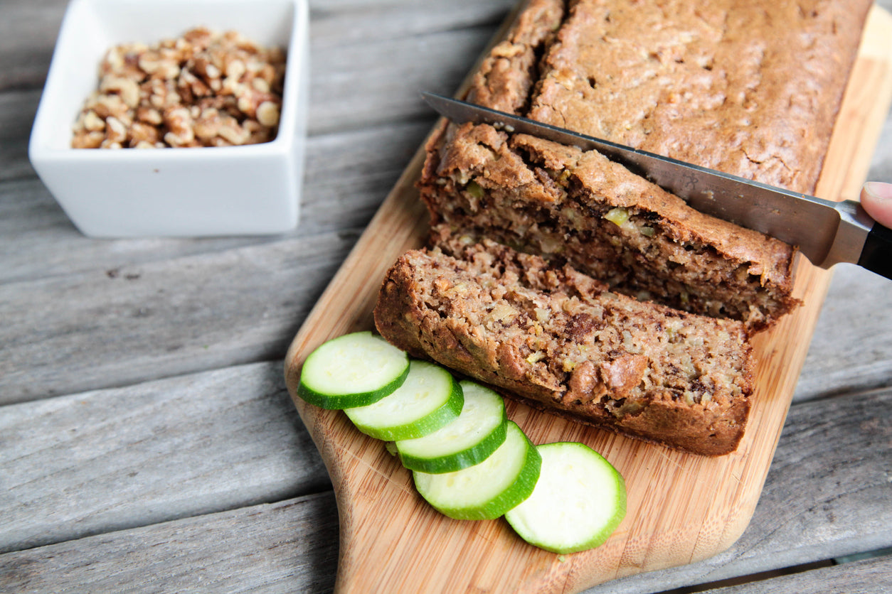 Zucchini bread being cut as it sits on a wooden cutting board. Slices of zucchini lay next to it, bright green. A bowl of crushed walnuts in the background.